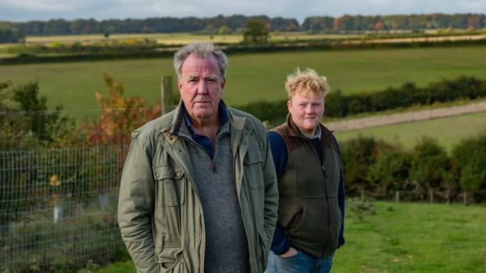 Two white men stand in the foreground of green farmland. The elder white man stares into the camera, while the younger of the two smirks at him