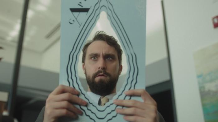 Corporate TV Show: A man sitting in a drab office looks through a hole in a piece of paper; he looks horrified. 
