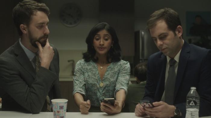 Corporate TV Show: Two men and a woman sit in an office breakroom; one o the men and the woman are looking at their phones. 