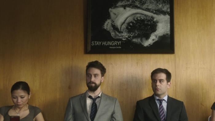 Corporate TV Show: Two men and a woman sit in a line on a couch; a corporate-type poster reading "Stay Hungry" appeals on the wood-paneling wall behind them. 