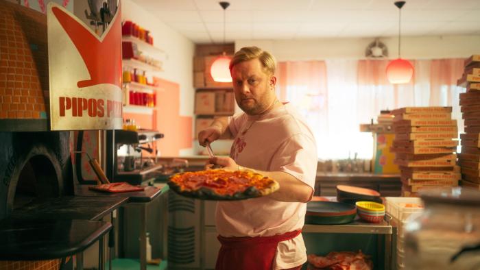 Crap Happens TV show screenshot 1: A man in a pizzeria holds a pizza near an oven, with pizza boxes stacked behind him.