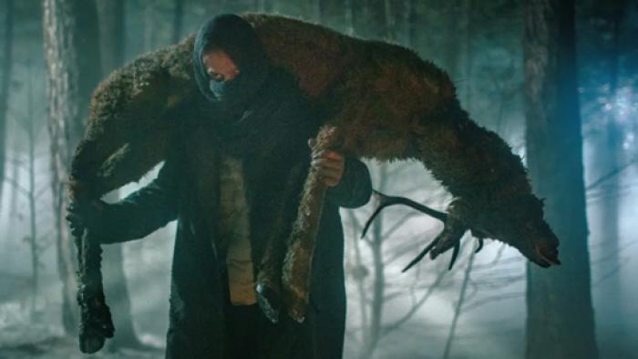 Creature TV show screenshot: A man stands in the snowy forest with a dead deer over his shoulders.