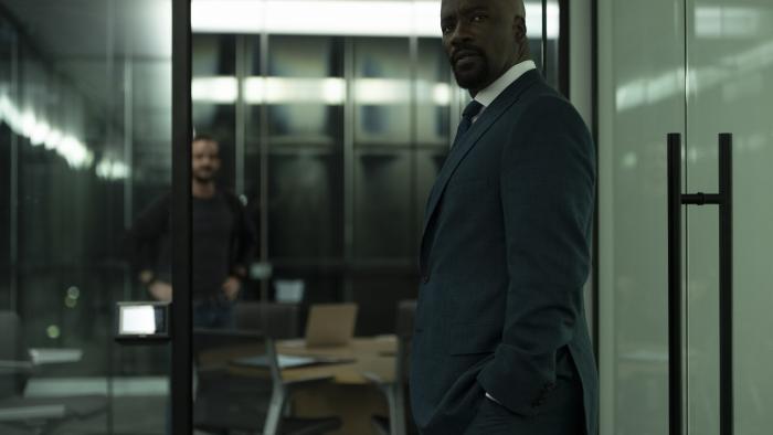 Cupertino TV show screenshot 3: Mike Colter stands in the doorway of a glass office. He wears a dark blue suit, dark tie, and white collared shirt.