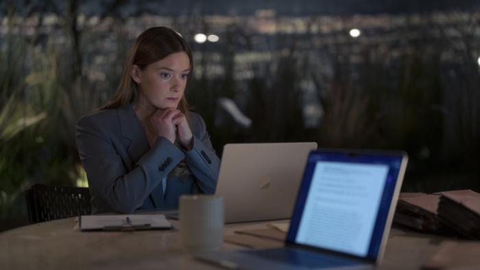 Cupertino TV show screenshot 1: Rachel Keller sits at a table with laptops on it. She wears a blue blazer and a white shirt.