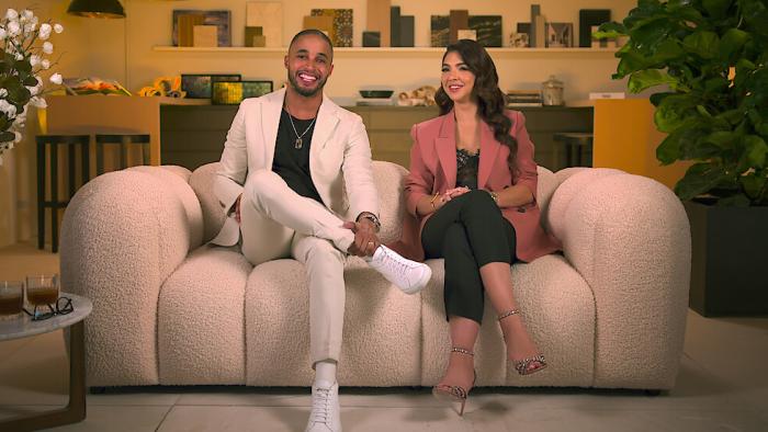 The two stars of Designing Miami sit on a puffy couch, smiling at the camera while conversing.