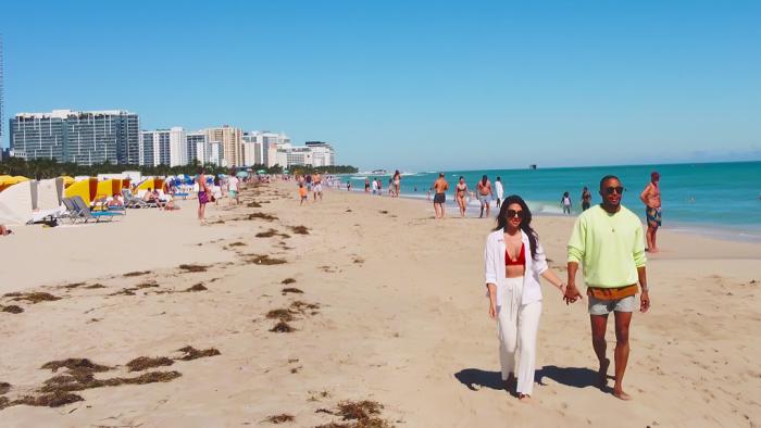 A man and woman walking on the sand at a sunny beach with people milling about in the background. The couple are holding hands and wearing sunglasses.