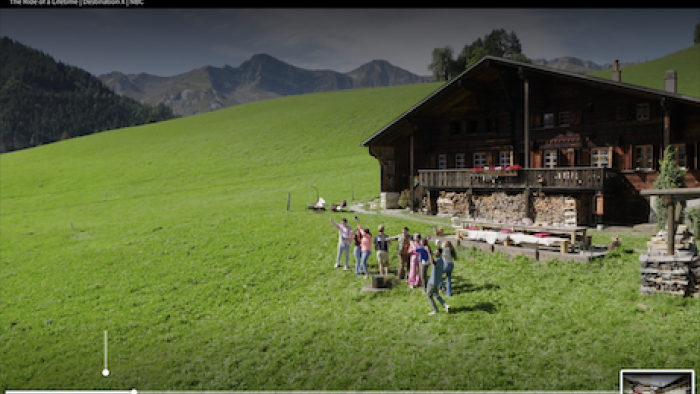 Destination X TV show screenshot 4: Contestants stand on grass nnext to wood house overlooked by snowy mountains.