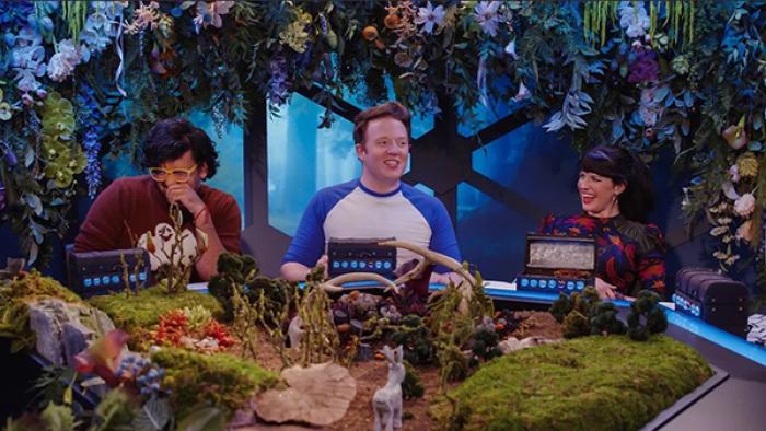 Dimension 20 TV show screenshot 2: Omar Najam, Brennan Lee Mulligan, and Emily Axford sit at the game table, framed with flowers and leaves