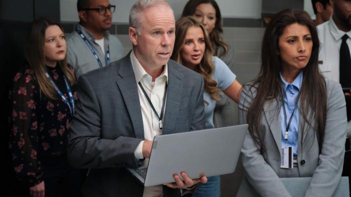 DMV TV show screenshot 3: (L-R) Gerry Dee and Reshma Shetty stand in a crowded bathroom. Dee holds a silver laptop.