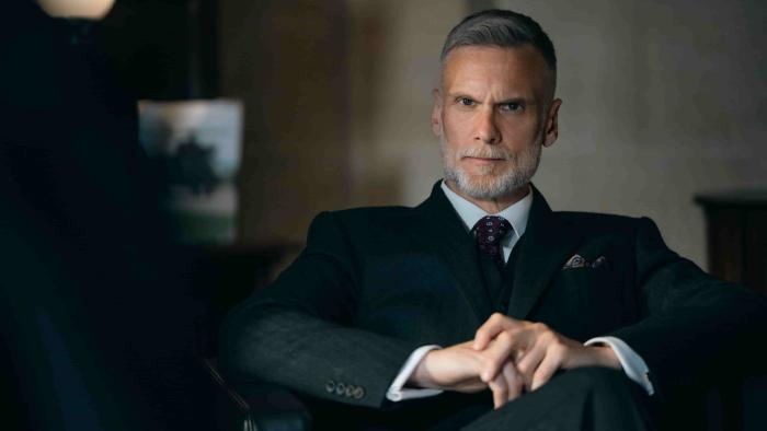 Down Cemetery Road screenshot 3: Darren Boyd sits cross legged in a leather chair. He wears a dark suit, burgundy patterned tie, and a pocket square.