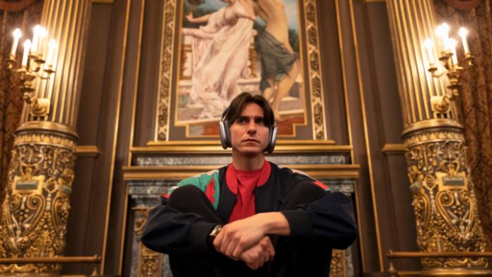 Étoile TV show screenshot 3: Tobias sits in an elegantly appointed room with elaborate carved walls, wearing headphones and making a disgusted face. 