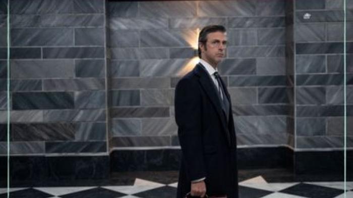 Kim Rossi Stuart stands in the safe room, wearing a long coat and holding a briefcase.