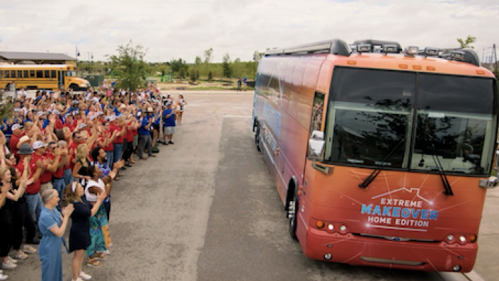 Extreme Makeover: Home Edition (2025) screenshot 3: Crowd dressed in red and blue stand and cheer by side of red Extreme Makeover bus.