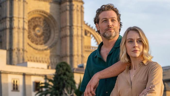 A White man rests his arm on a White woman's shoulder in front of a cathedral