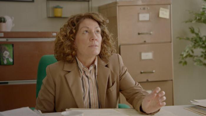 Fisk TV show screenshot 1: Kitty Flanagan sits in an office, in a green chair. She wears a brown suit.