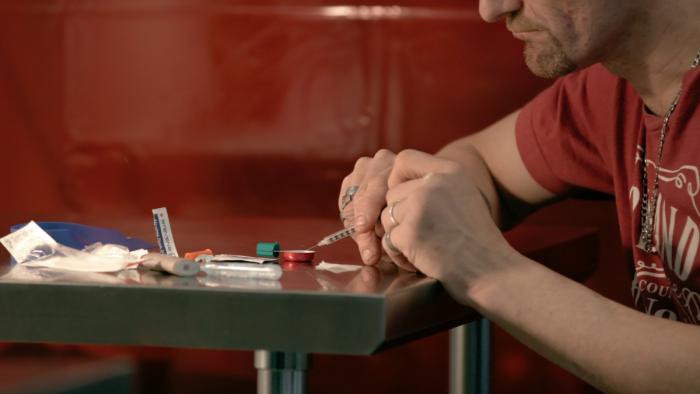 Follow This Series: A man sticks a syringe into a substance mixed up in a metal dish; on the table in front of him are a lighter and a bag of white powder. 