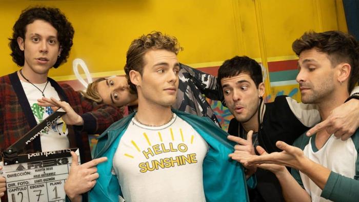 Freeks TV show screenshot 1: Three band members stand admiring their bandmates t-shirt, while another stares ahead holding a clapboard; yellow background. 