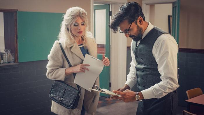 Barbara wears a beige coat, holding a script, while Dennis stands next to her looking down at a clipboard