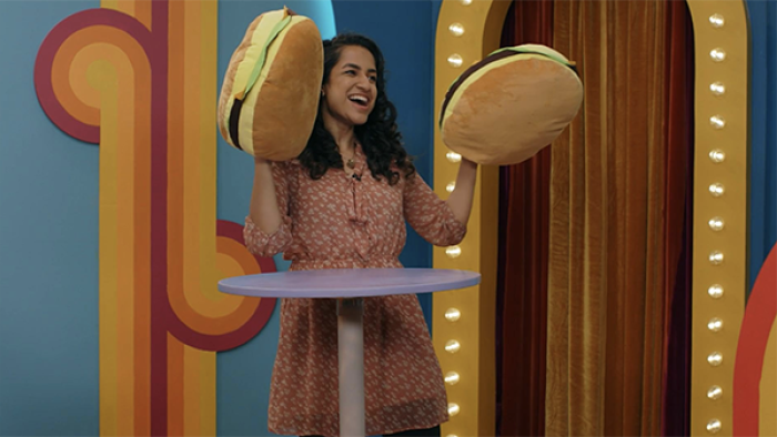 Game Changer TV show screenshot 2: Rekha Shankar wears a coral-colored dress and golds up her hands, which are covered with big plushy hamburgers