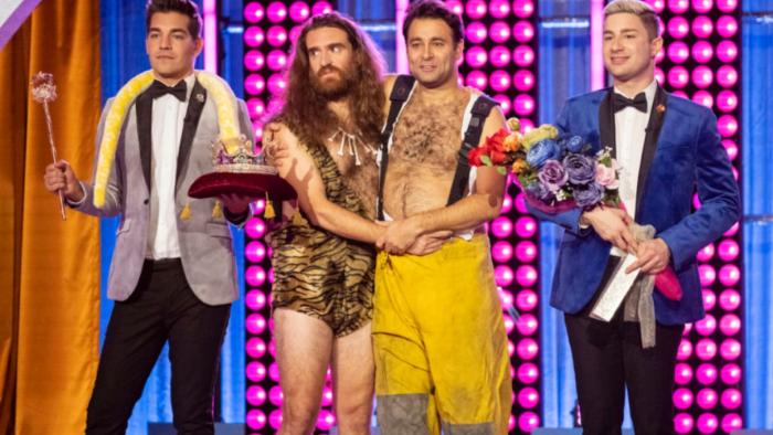 Gayme Show TV show: Four White men stand on stage; the middle two in caveman and firefighter costumes hold each other