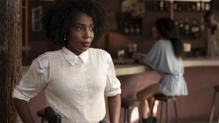 Tamara Lawrence looks off to the side as Chyna McQueen sits behind her. She wears a white collared shirt, beige vest, and gun.