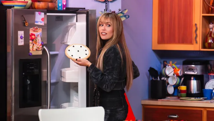 Hannah Montana 20th Anniversary Special screenshot 2: Miley Cyrus in Hannah Montana's kitchen holds a pie in her hand with the fridge door open.