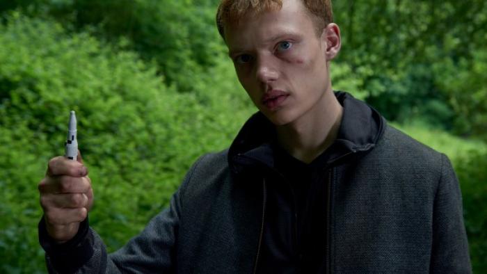 Hard Sun Series: A young man with intense eyes and a grim expression holds up a marker in front of a green leafy landscape. 