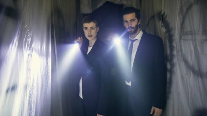 Hard Sun Series: A man and a woman, both in dark business suits, each shine their flashlights on something off-camera in front of a creepy background covered with clear sheeting and graffiti. 