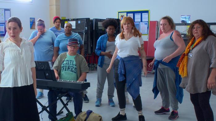 Hard Cell TV show screenshot 1: A group shot of various characters standing around what looks like a rec room in a prison. One sits at al electronic keyboard.