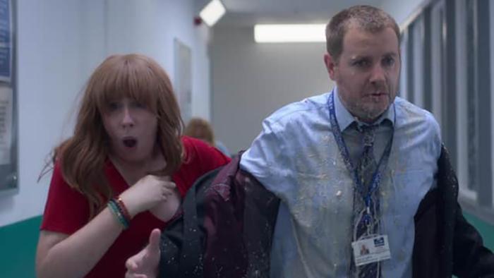 Hard Cell TV show screenshot 2: Two characters walk down a hallway lit by fluorescent lights. On the left, a redheaded woman in a red top clutches her chest, and on the right, a man with short hair and a light blue shirt looks bewildered. He is covered in some kind of gloopy, wet substance.