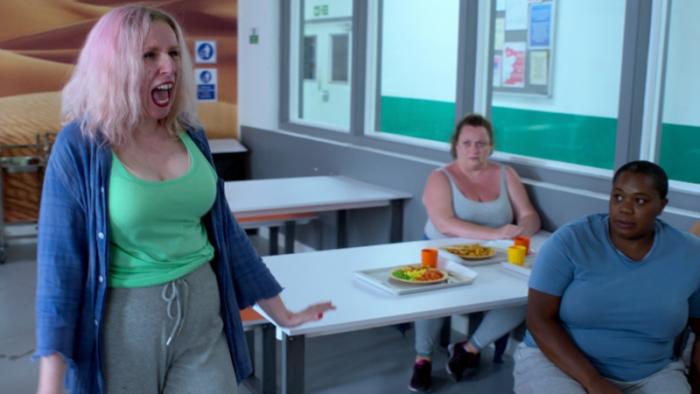 Hard Cell TV show screenshot 3: A pink-haired woman in a green shirt stands in a cafeteria, shouting, as two seated women look on.