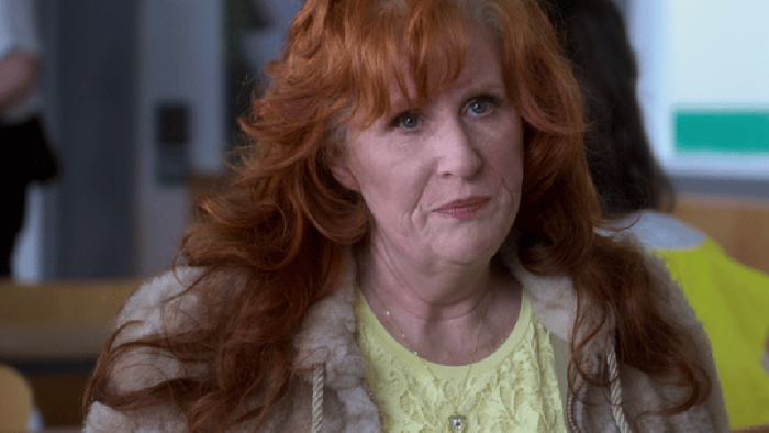 Hard Cell TV show screenshot 4: A woman with red hair and white roots is pictured sitting down with an annoyed expression on her face.