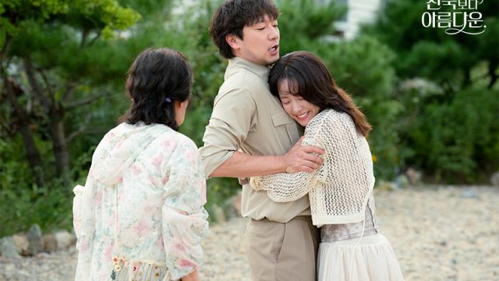 A young man in khaki pants and matching shirt is embraced by a young woman but the man is trying to pull away. An older woman looks on and watches.