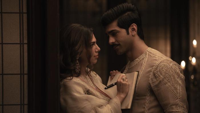 Sharmin Segal holds a book and pen against Taha Shah Badussha’s chest while gazing into each other’s eyes.
