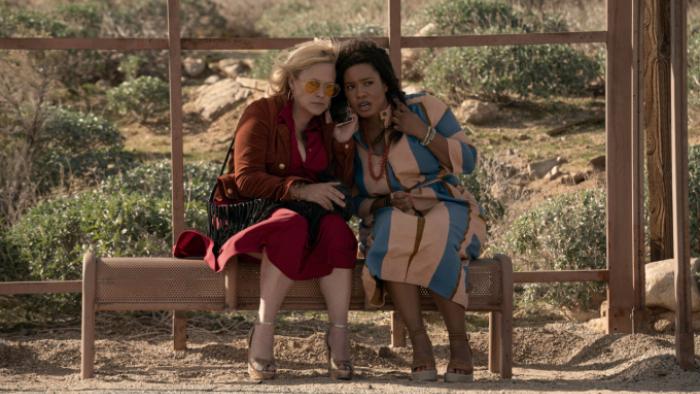 High Desert: Peggy and Carol sit together on a bench in front of a window showing a desert background. Their faces are close together as they listen to something dismaying on a cell phone. 