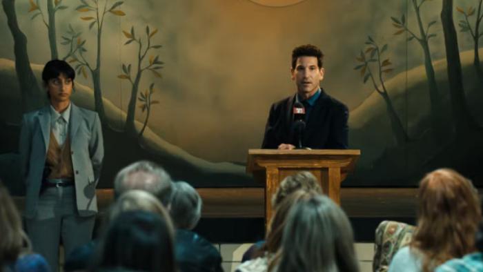 His & Hers TV show screenshot 3: Richard stands at a podium in front of a small audience while Priya is off to one side