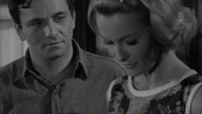 A man with dark hair looks at a blonde woman expectantly, she is glancing down.