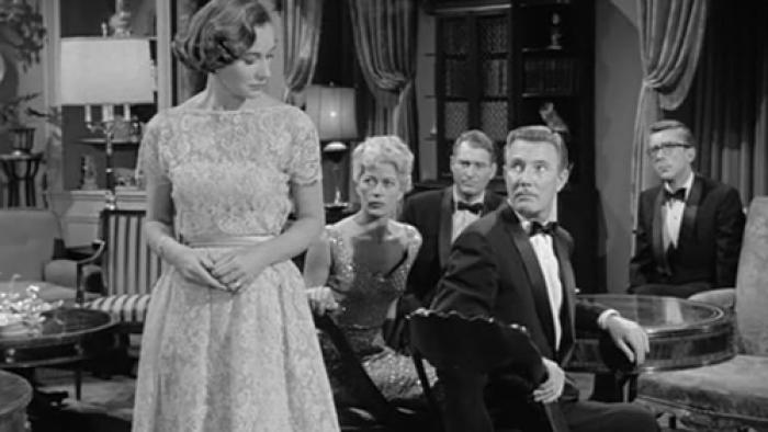 Two women in formal dresses and three men in tuxedos sit and stand in a well-appointed room with velvet curtains.