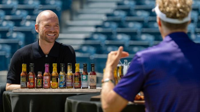 Hot Ones: Extra Heat TV show screenshot 3: Sean Evans sits smiling in front of a row of hot sauces, facing Will Ferrell