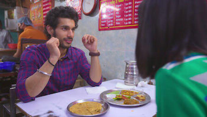 A South Asian man in a plaid shirt talks to a South Asian woman wearing green as they sit at a table in a restaurant.