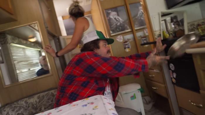 Just a Dash TV show screenshot 2: Matheson leans to catch a pan sliding off the stove. He wears a flannel shirt, trucker hat, and white tee.