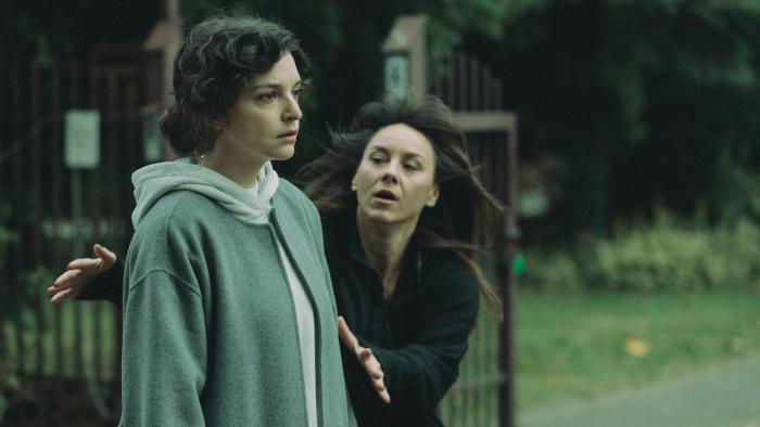 One woman wearing all black rushes to another woman who wears drab grey and looks dazed and exhausted. They are both outside, it looks dark and cold