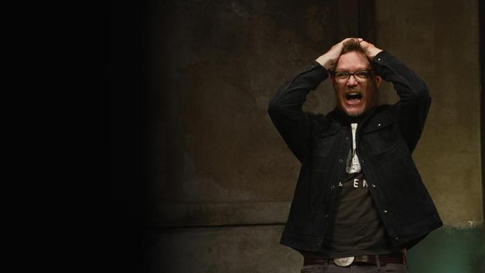 In a darkened corner, host Matthew Lillard stands wearing dark clothing and glasses and with his hand on his head expressing emotion.
