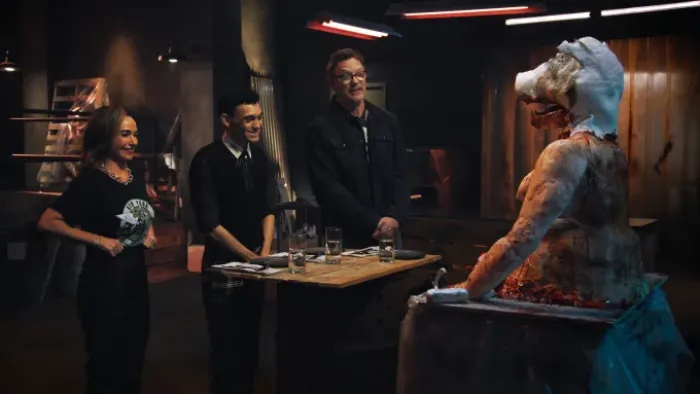Darkened room with the three show hosts smiling uncertainly and grimacing at a large cake in the shape of a fleshy pig-like being.