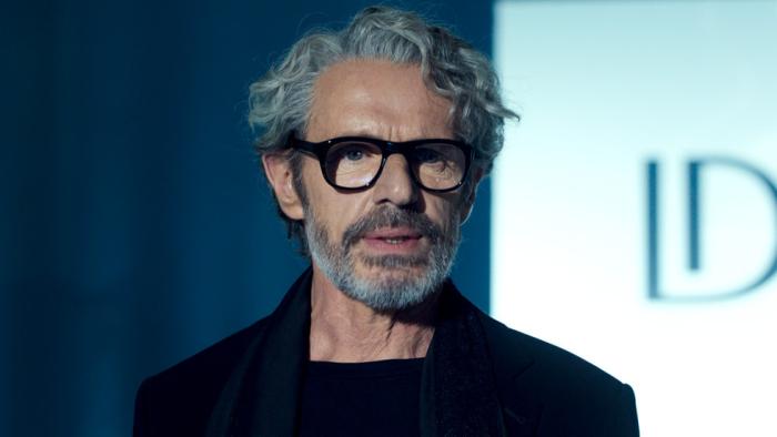 La Maison TV show screenshot 1Lambert Wilson stands in the spotlight. He wears thick rimmed black glasses.
