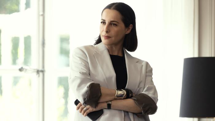 La Maison TV show screenshot 3: Amira Casar wears a white suit jacket, a black shirt, gold bangles, and holds a phone.