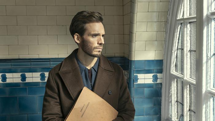 Joel "Laz" Lazarus is shown holding a brown folder to his chest as he stares out a window with a glum expression on his face