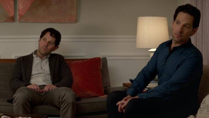 Living with Yourself Series: The two Mileses sit together in a living room, craning their necks to see something off-screen. 