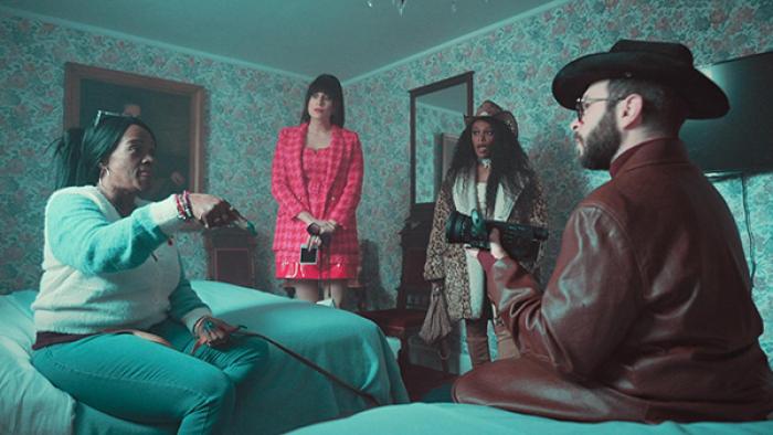 Living for the Dead TV show screenshot 2: A woman sits on a bed in a bedroom with floral wallpaper, animatedly speaking with three of the team's investigators. One of the team members listens on with her jaw dropped.