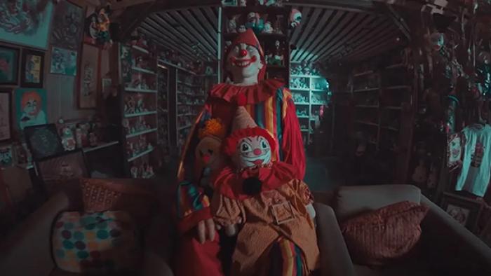 Living for the Dead TV show screenshot 3: A cluster of clown toys is in the foreground of a dark room filled with two chairs and an abundance of clown paintings, figures, and other collectibles.